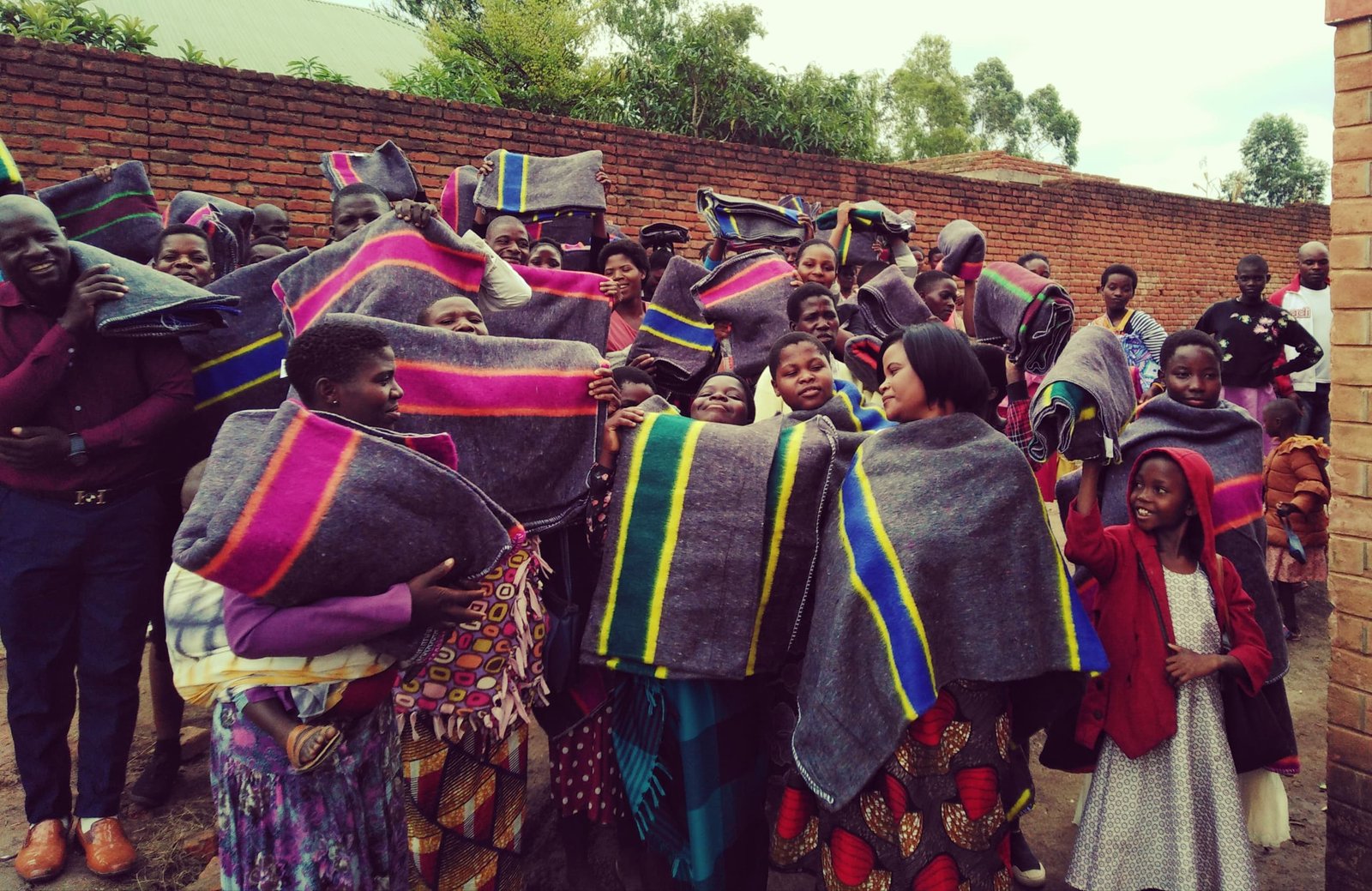 A group stands beside a brick wall, holding dark blankets with bright stripes.