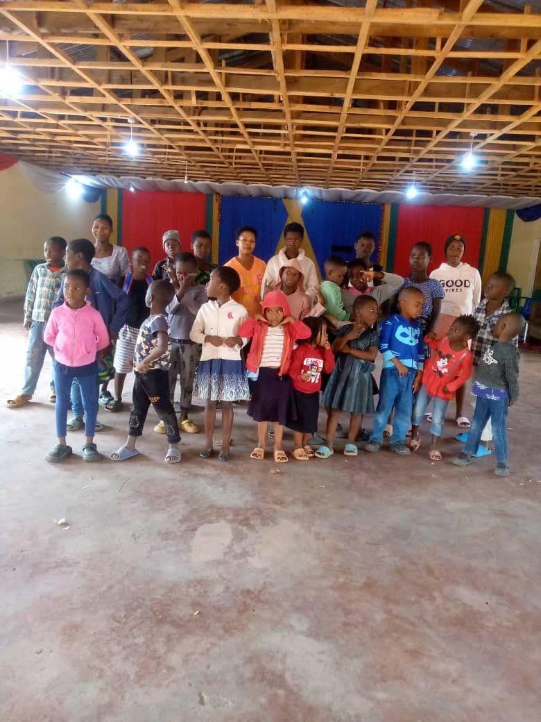 Children and adults stand at the front of a hall beneath an exposed timber roof.