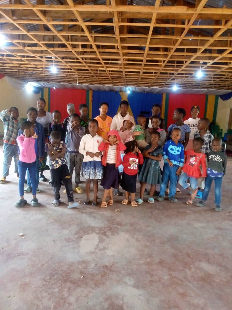 Children and adults pose together in a hall with red, white, and blue drapery.
