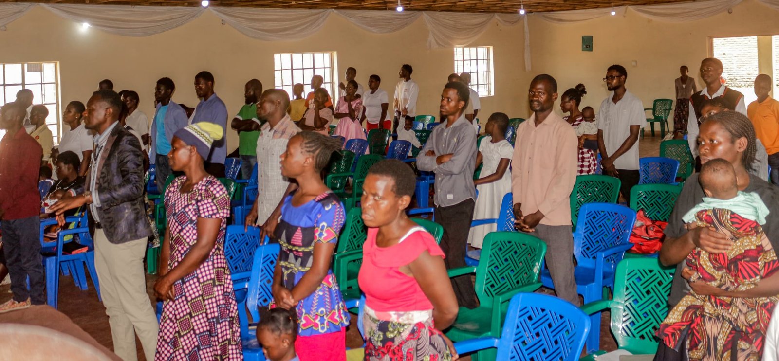 Adults and children stand among blue and green chairs inside a church hall.
