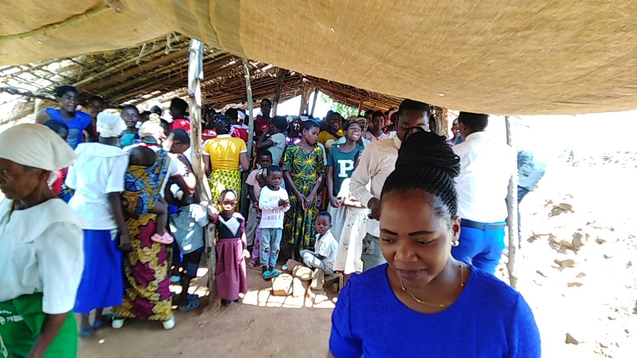 A woman in blue in the foreground of a gathering beneath a thatched roof.