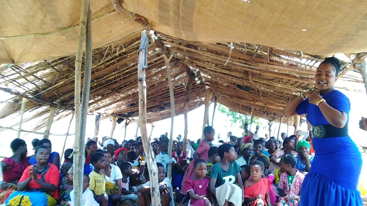 A woman speaks with a microphone beside a seated congregation under a thatched roof.