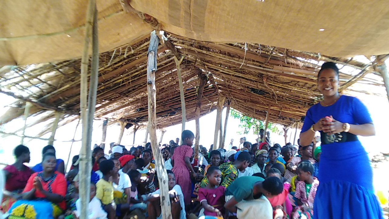 Another moment of a woman speaking beside the congregation under a thatched roof.