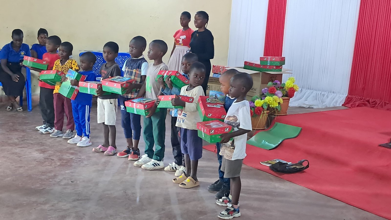 Younger children hold red and green boxes at the front of a hall.