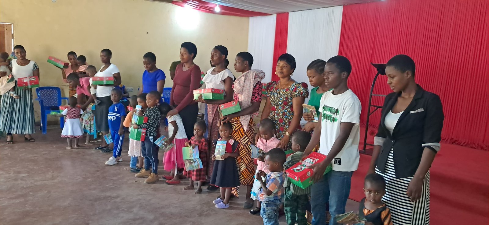 Adults and children stand together holding red and green boxes.